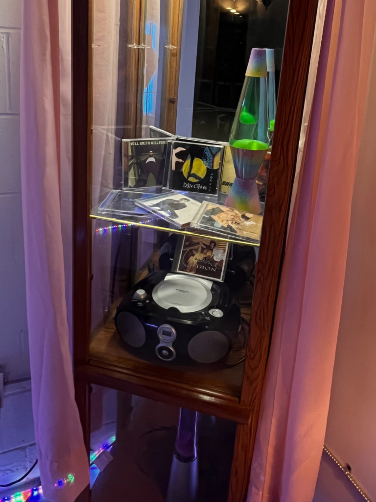 image of a glass, mirrored cabinet with Y2K themed items inside. a lava lamp, a CD player, and some CD's can be seen inside the cabinet, and the entire cabinet is encased in a pink, sheer princess circular drape.