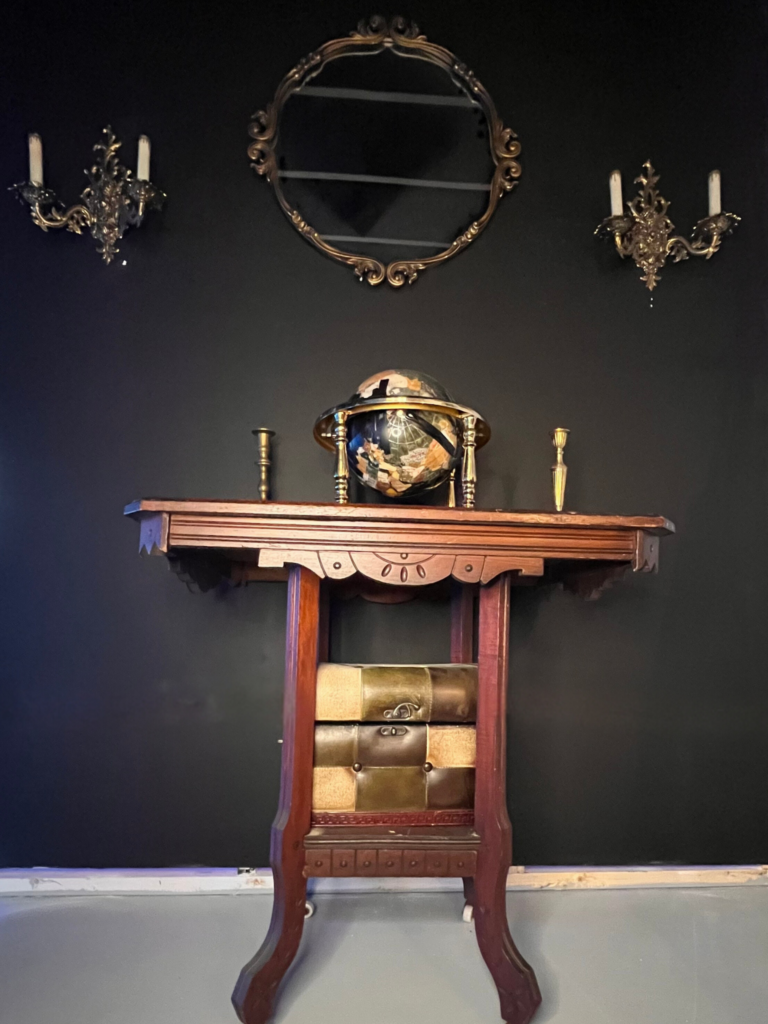image of a wall in the Dark Academia self-photography set. This wall features an antique dark, wooden side table with an antique black and gold globe, old candle stick holders, and a treasure chest on it. Above the table is an antique, round mirror, with candle stick sconces on either side of it.