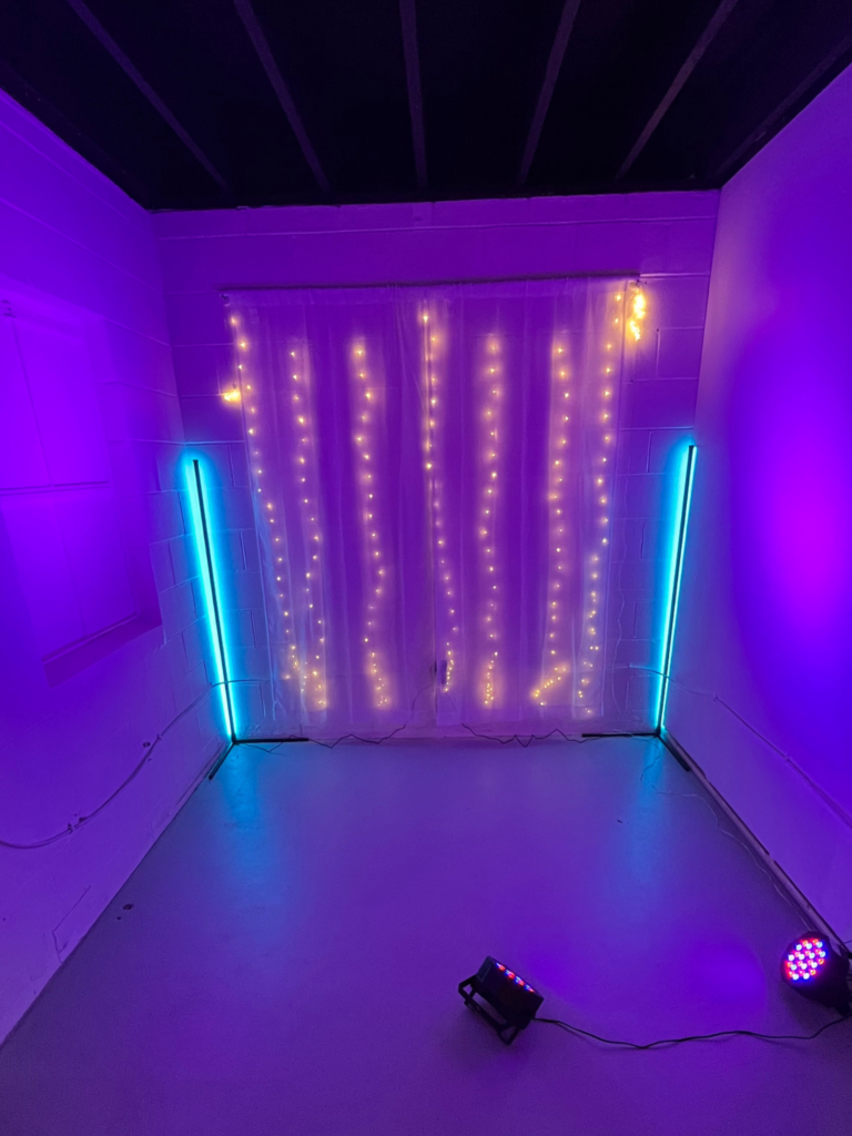 image of a self-photography station featuring blue and purple lighting, a white sheer curtain backdrop with warm fairy lights shining through.
